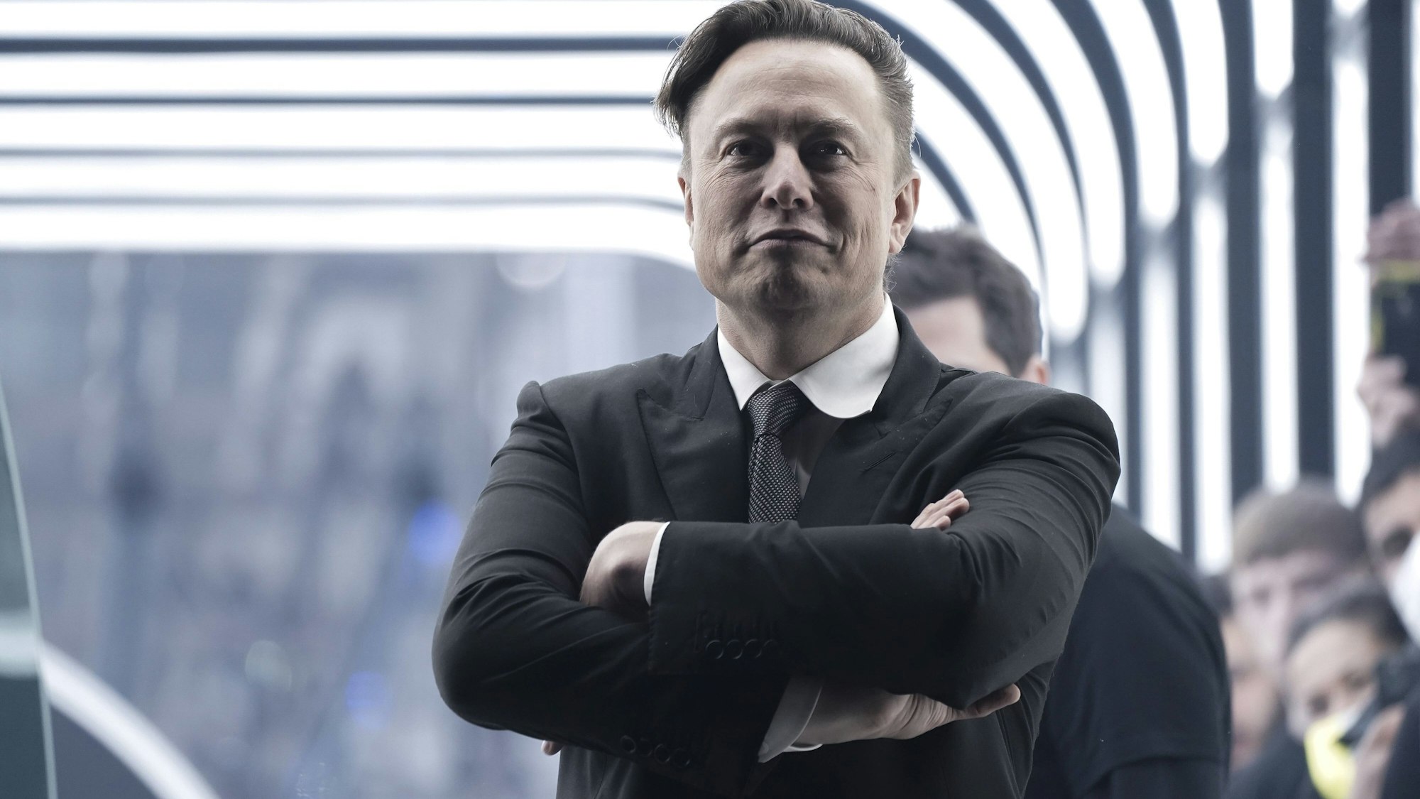 Elon Musk steht mit verschränkten Armen in einer Werkshalle.