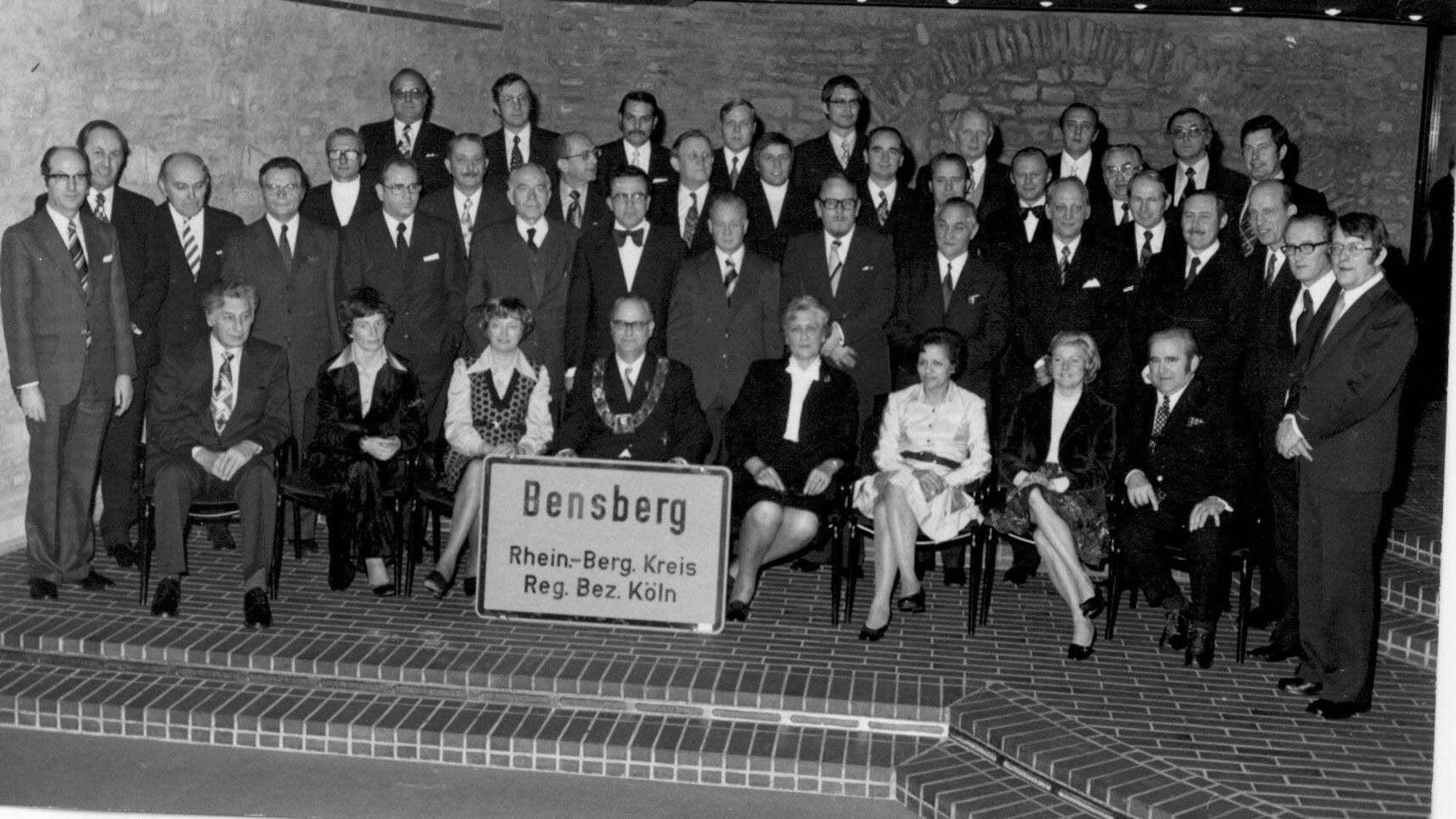 Die Mitglieder des Stadtrates der Stadt Bensberg stehen am 18. Dezember 1974 während ihrer letzten Sitzung mit einem Bensberger Ortsschild im Ratssaal des Bensberger Rathauses.