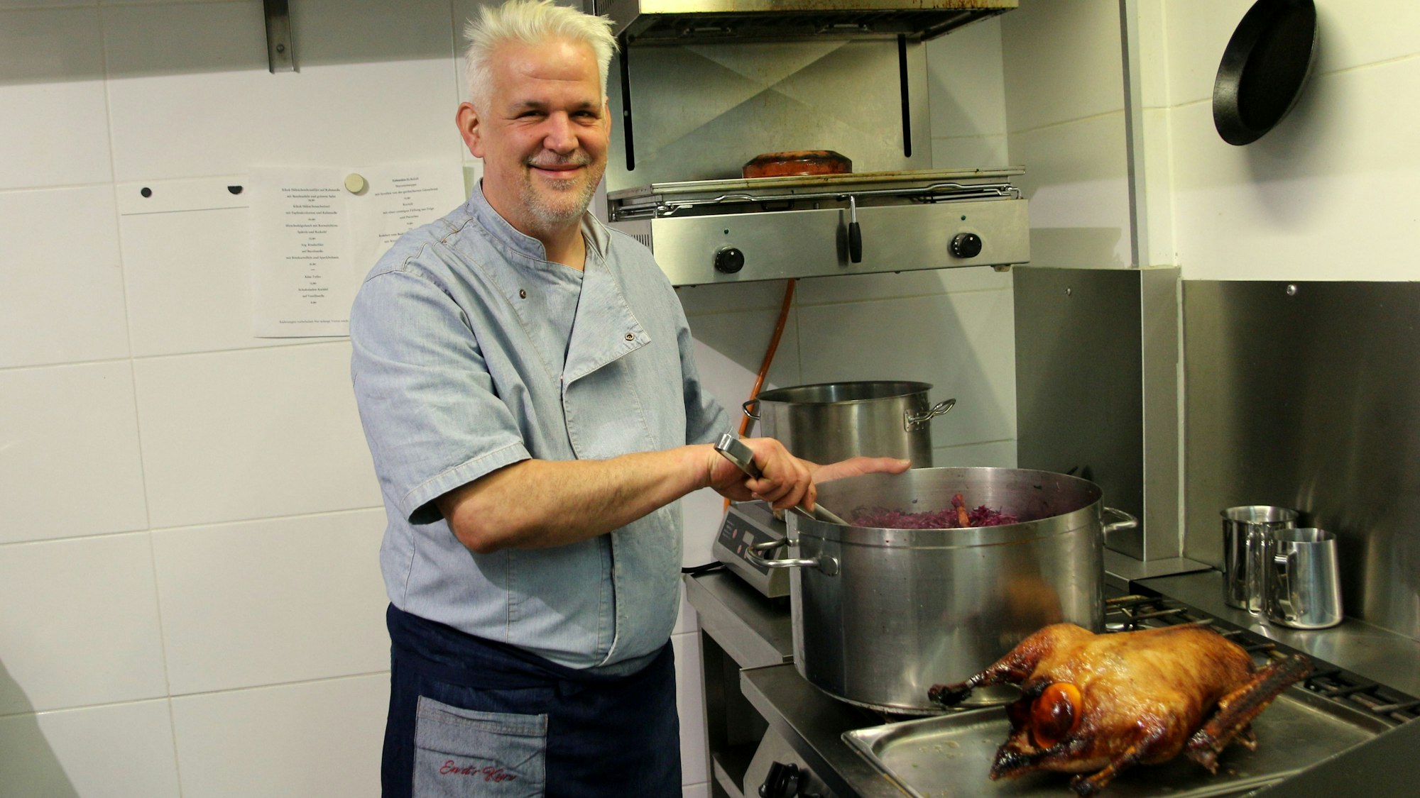 Auf dem Bild ist ein Koch an einem großen Topf zu sehen, neben ihm ein großer gebratener Vogel.