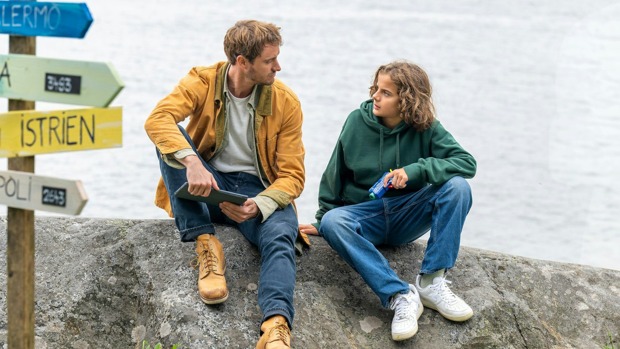 Filmszene aus „Das Flüstern der Pferde“: Nils (Frédéric Brossier) und Peppe (Asterios Rose) sitzen an einem Gewässer auf einem Felsen und blicken sich an.