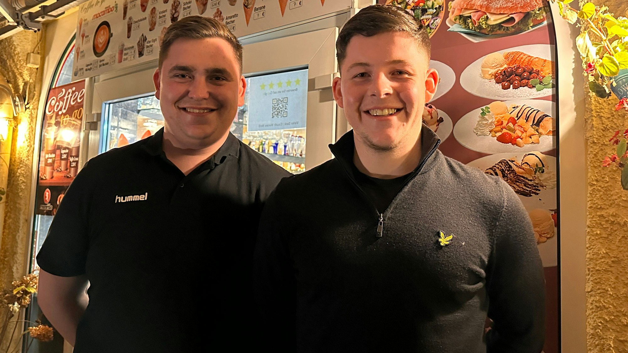 Zwei junge Männer, die schwarze Shirts tragen, stehen lächelnd vor einem Eiscafé.
