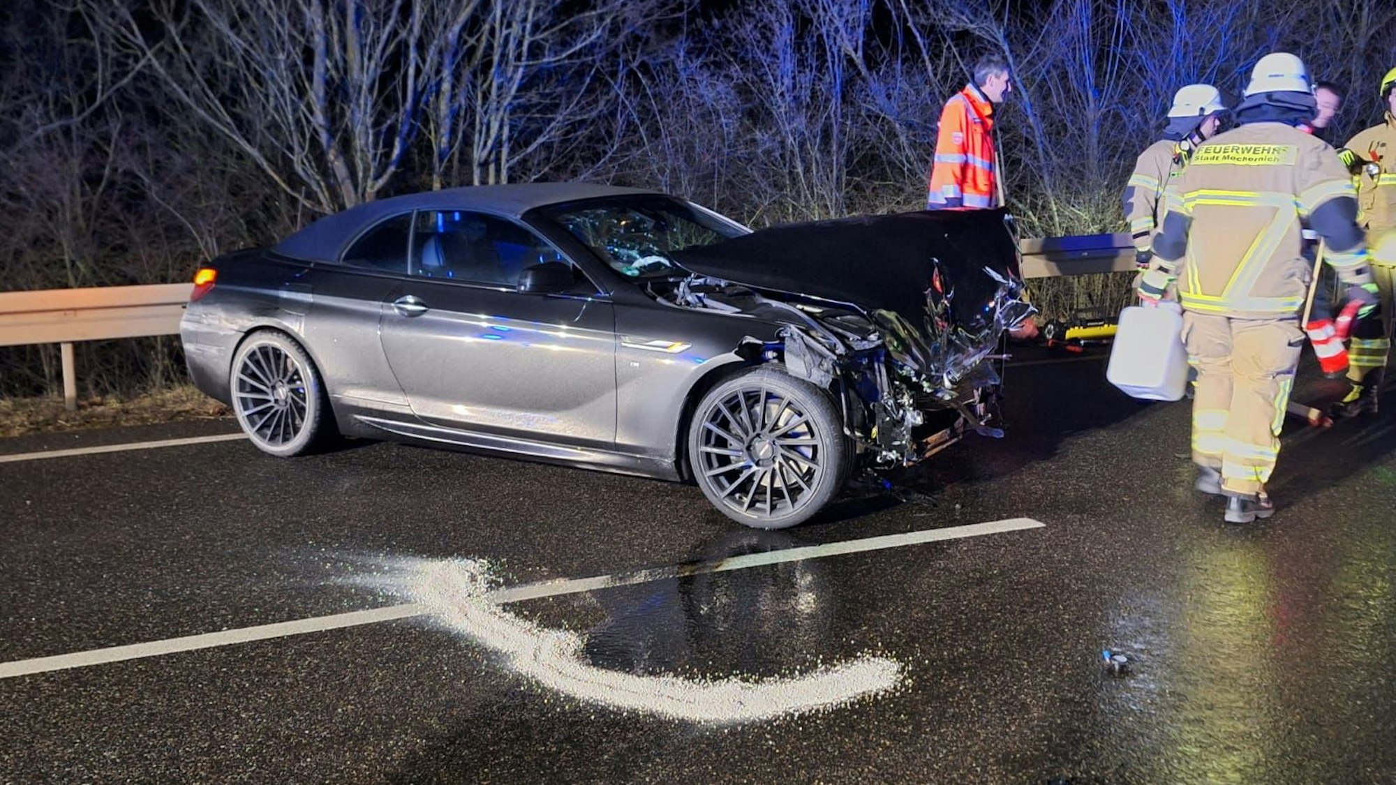 Ein silbergrauer Wagen steht mit zerstörter Front auf quer auf einer Straße. Daneben sieht man Feuerwehrleute, die die Fahrbahn abstreuen.