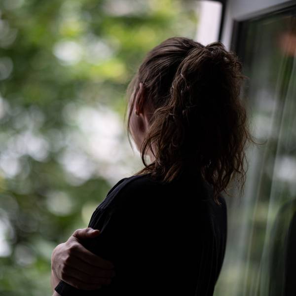 Eine Frau mit Pferdeschwanz steht mit dem Rücken zum Betrachther an einem geöffneten Fenster und schaut hinaus.