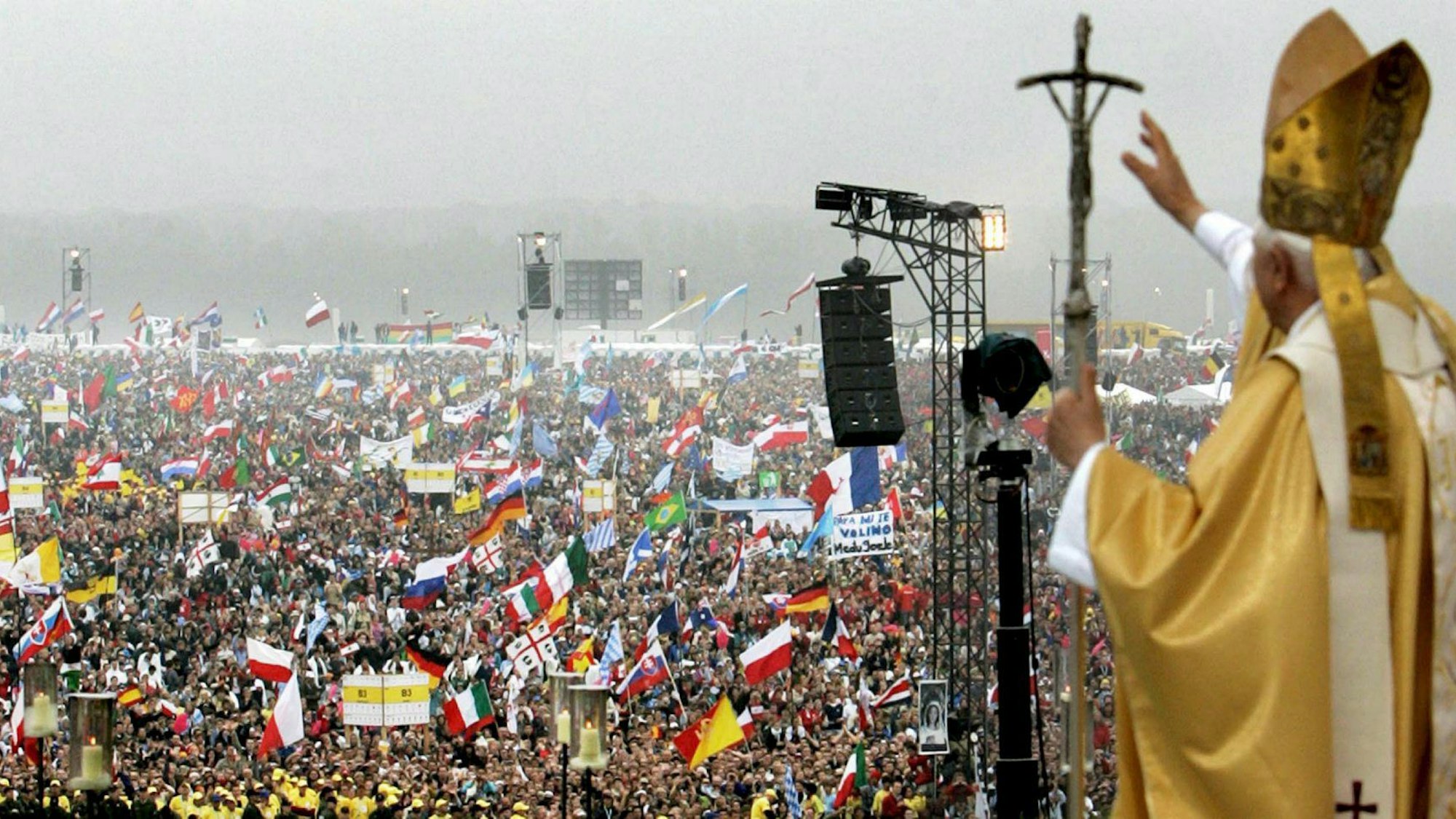 Papst Benedikt XVI. winkt einer Menschenmenge, in der zahlreiche Fahnen geschwenkt werden, von erhöhter Stelle aus zu.