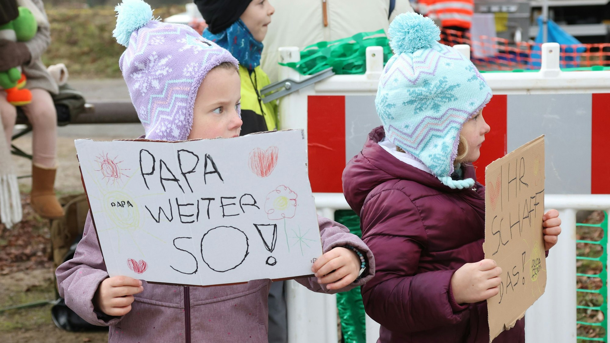 Zwei kleine Kinder mit Pudelmützen halten Pappschilder in den Händen. Darauf zu lesen steht "Papa, weiter so!" und "Ihr schafft das!".