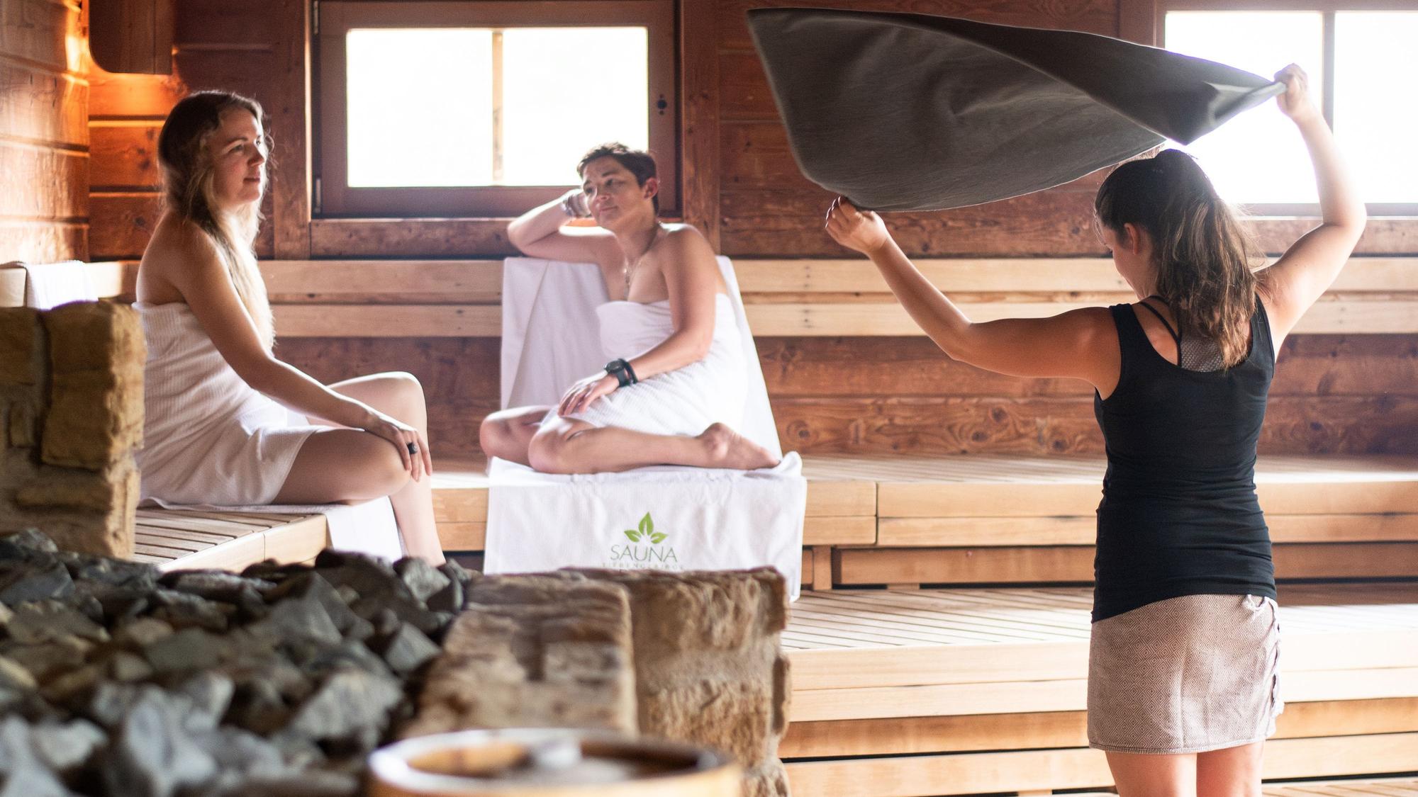 Zwei Frauen sitzen in der Sauna, eine Mitarbeiterin wedelt mit einem Handtuch nach dem Aufguss.