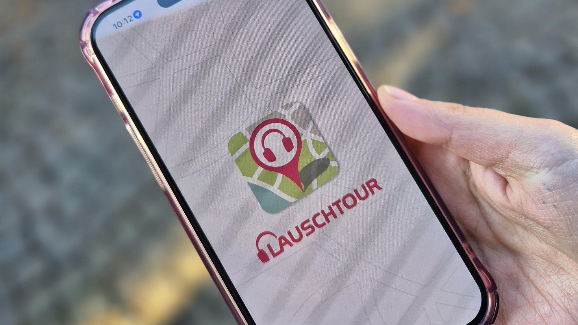 Eine Hand hält ein Smartphone fest, auf dessen Bildschirm die App Lauschtour zu sehen ist.