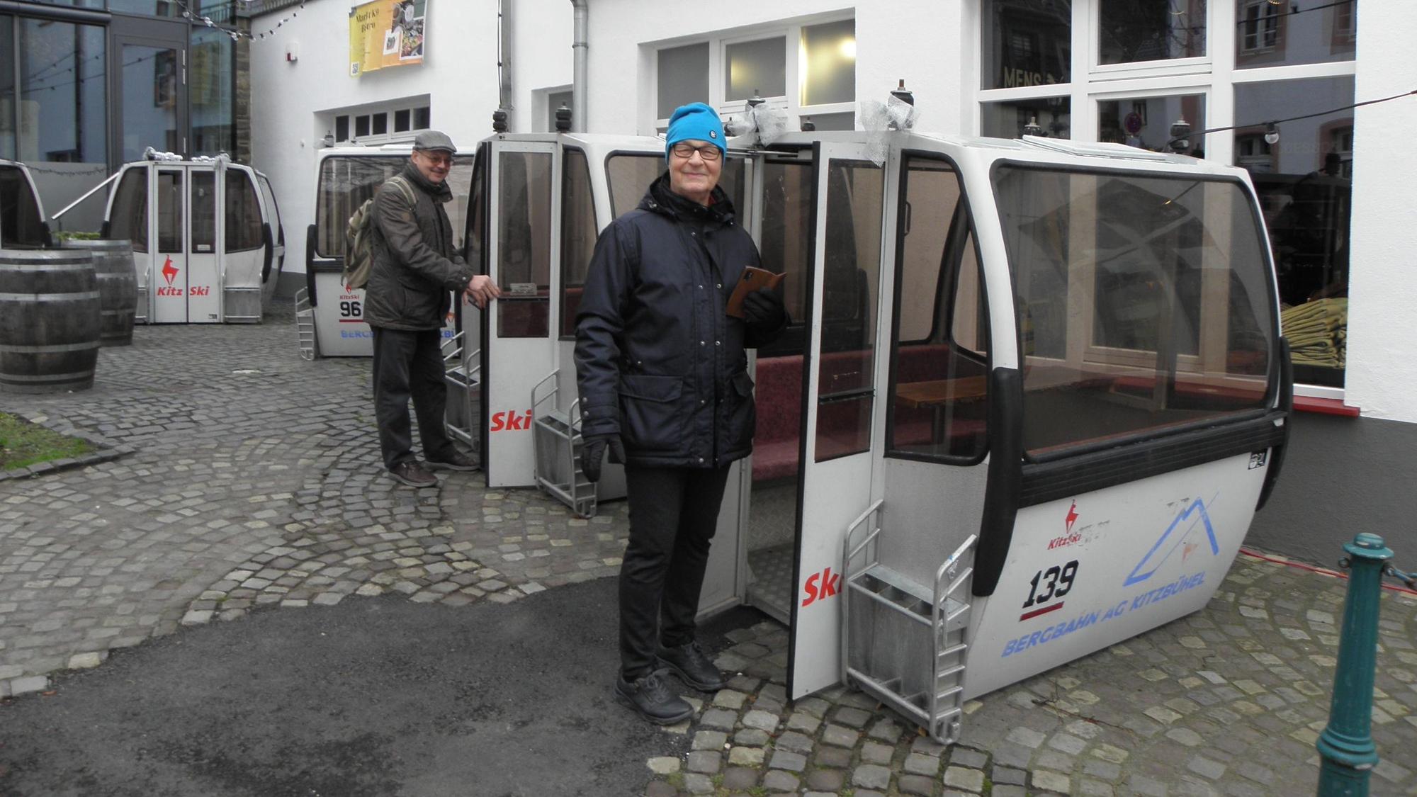 Mehrere Seilbahnkabinen stehen auf einem Parkplatz, ein Mann und eine Frau schauen sie interessiert an.