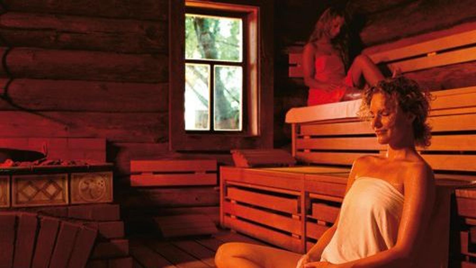 Zwei Frauen, die sich Handtücher umgewickelt haben, entspannen in einer Sauna, die rötlich beleuchtet wird.