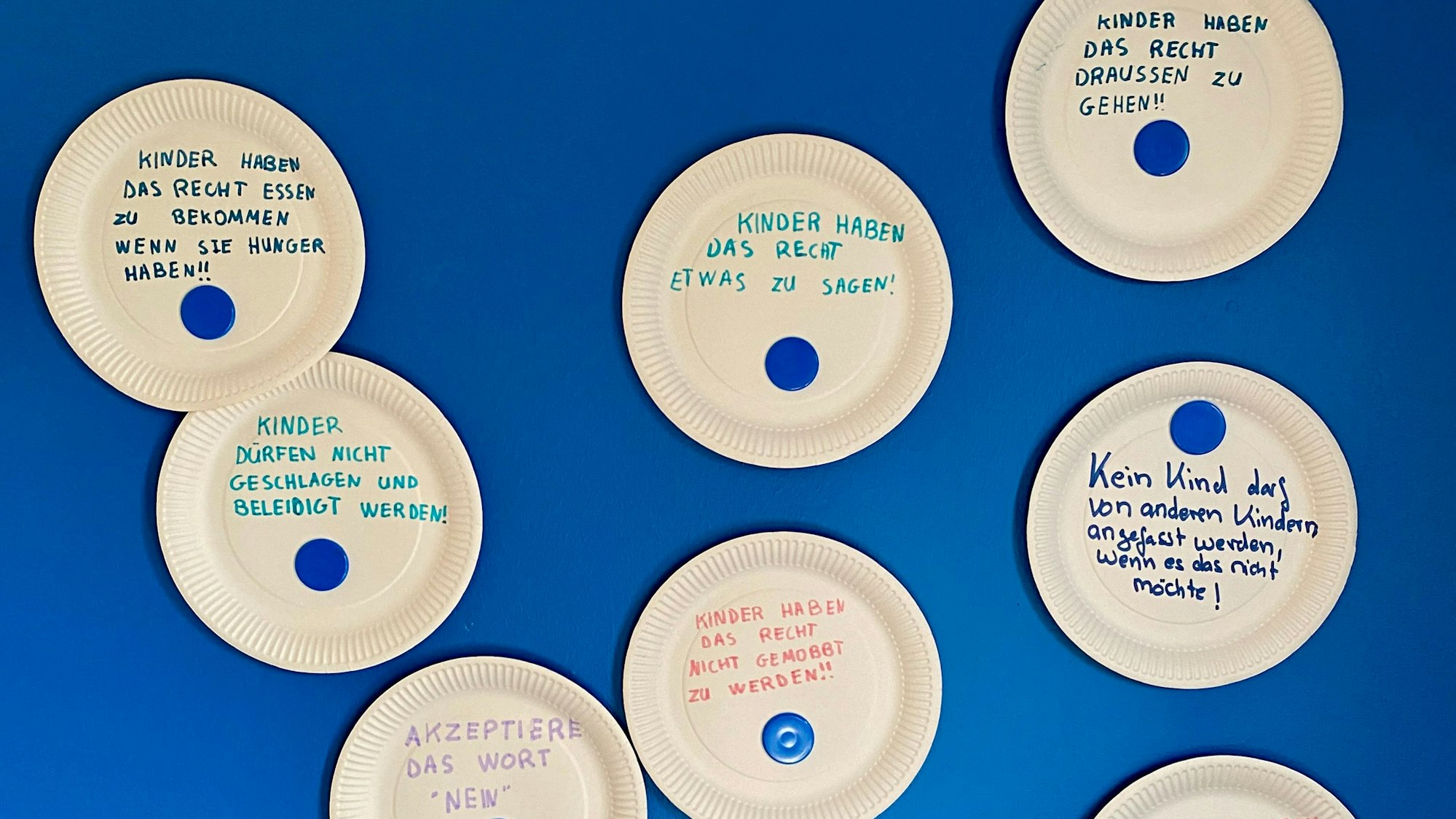 Pappteller hängen an einer blauen Wand. Mit bunten Filzstiften sind darauf einige Kinderrechte geschrieben.