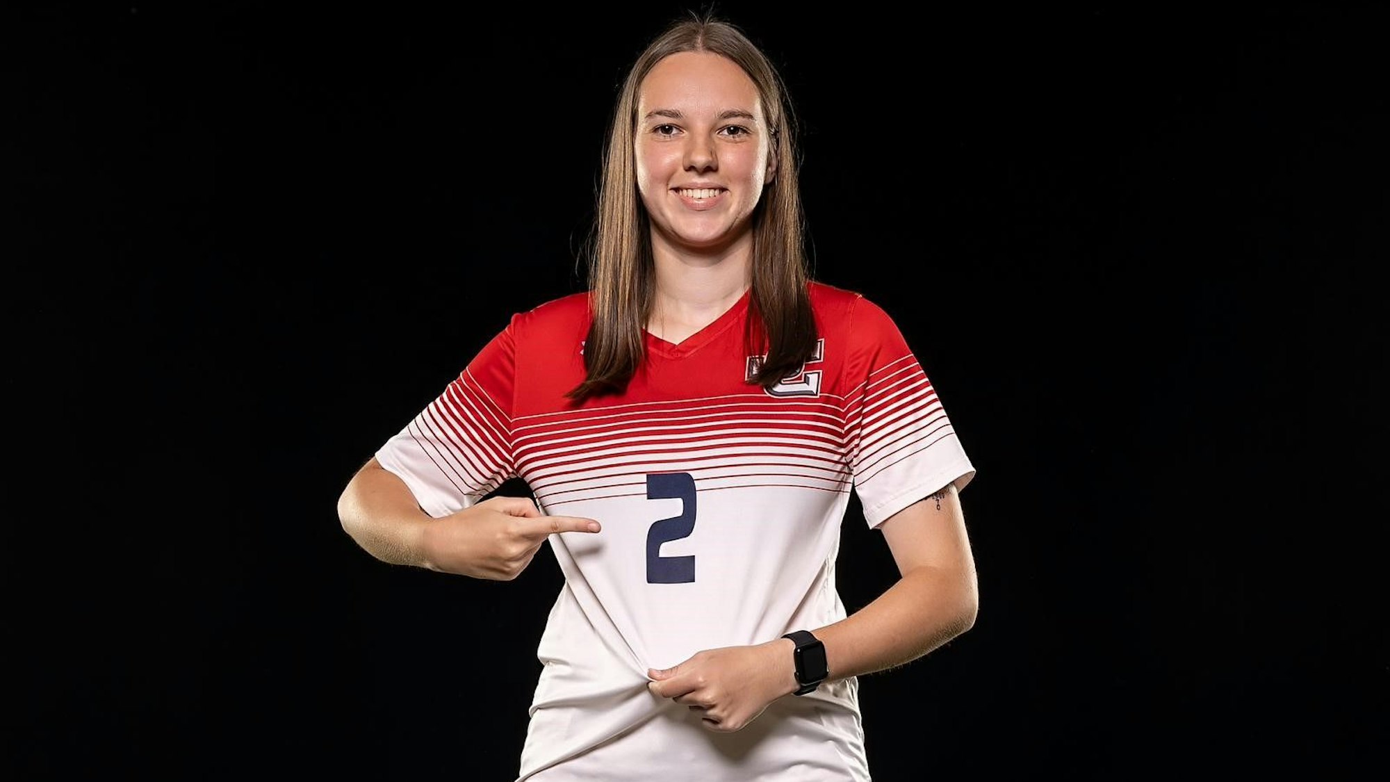 Lena Kücken trägt das Trikot mit der Nummer 2 des Jefferson College. Mit einem Finger zeigt sie auf die Nummer, mit der anderen Hand zieht sie das Trikot straff.