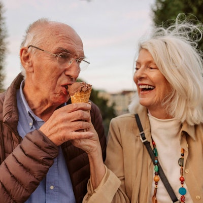 Eine ältere lachende Frau hält einem älteren Mann ein Eis an den Mund