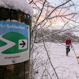 Eine Spaziergängerin mit Hund auf der verschneiten Eifelschleife Ahekapelle im Genfbachtal bei Nettersheim.
