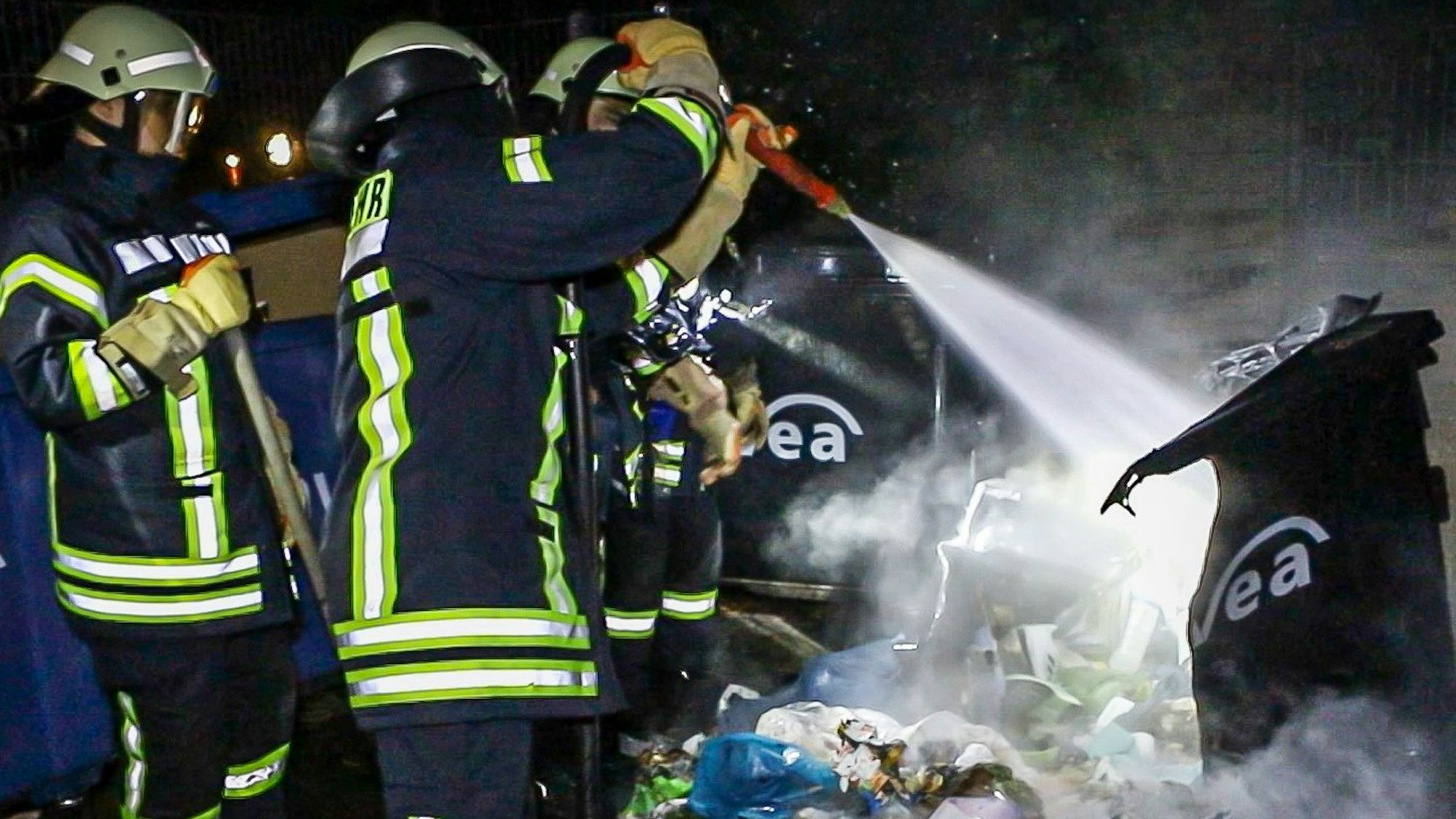 Feuerwehrleute löschen einen brennenden Müllcontainer.