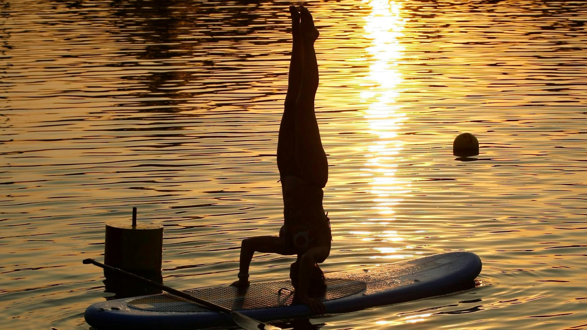 Eine Frau macht einen Kopfstand auf einem Stand Up Paddle Board auf dem Wasser im Sonnenuntergang.