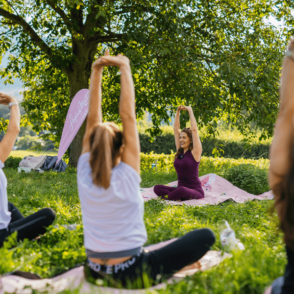 Die Yoga-Lehrerin leitet Teilnehmerinnen der Yogastunde während einer Yoga-Übung an. Sie sitzen bei gutem Wetter auf einer grünen Wiese.