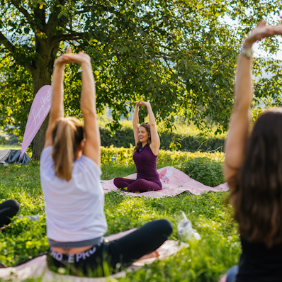 Die Yoga-Lehrerin leitet Teilnehmerinnen der Yogastunde während einer Yoga-Übung an. Sie sitzen bei gutem Wetter auf einer grünen Wiese.