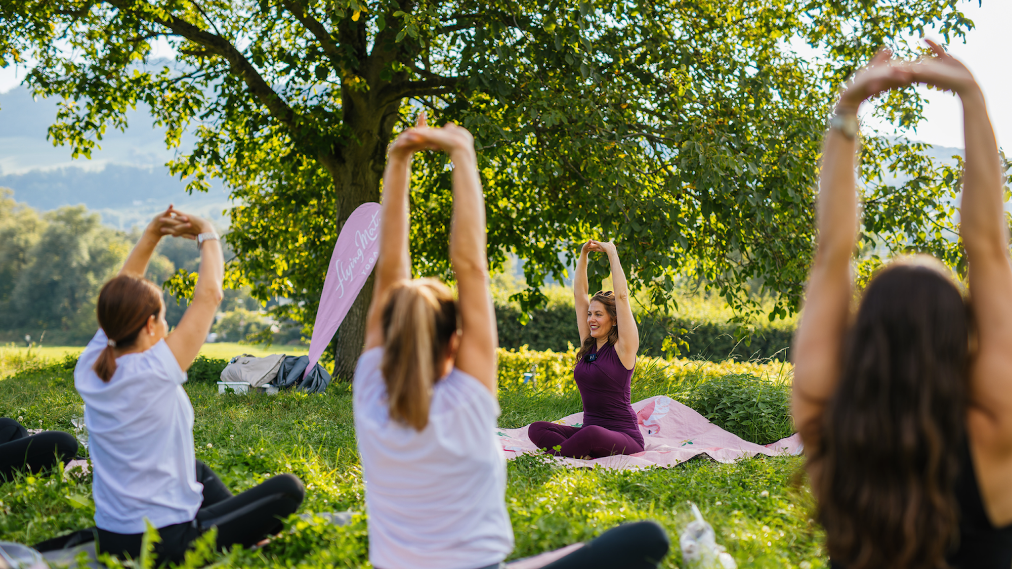 Die Yoga-Lehrerin leitet Teilnehmerinnen der Yogastunde während einer Yoga-Übung an. Sie sitzen bei gutem Wetter auf einer grünen Wiese.