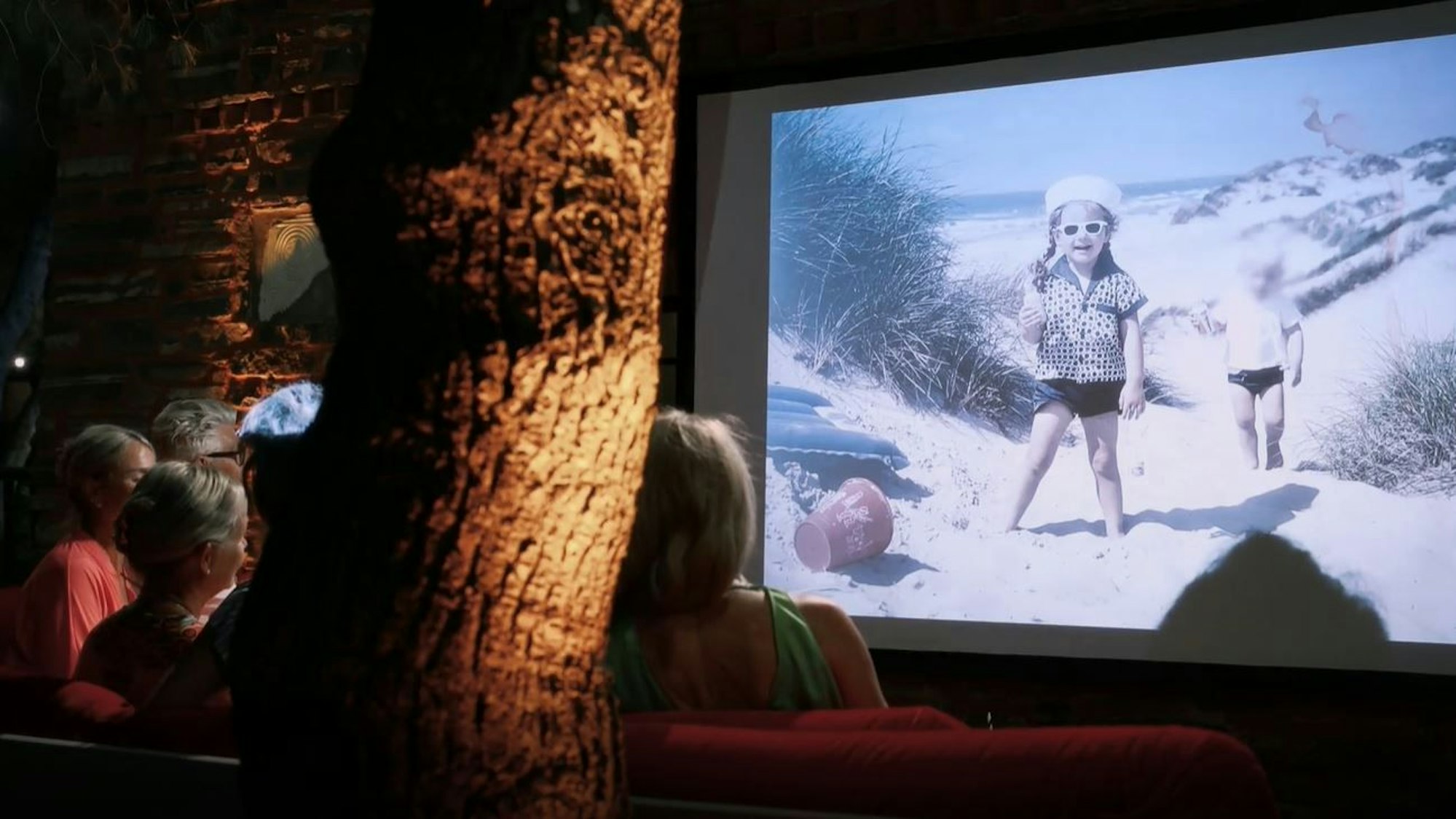 Sabine als Kind am Strand, mit Sonnenbrille auf der Leinwand im Freiluftkino. Links im Bild sieht man das rote Sofa, auf dem die Kandidatinnen und Franz sitzen.