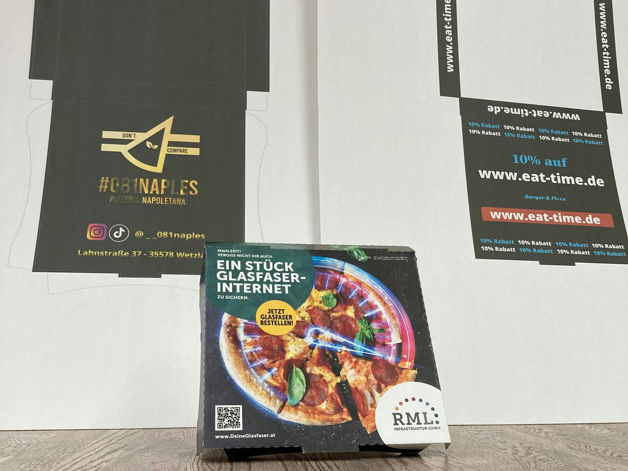 Ein Pizzakarton steht fertig gefaltet vor zwei Kartons, die die Innenseite zeigen.