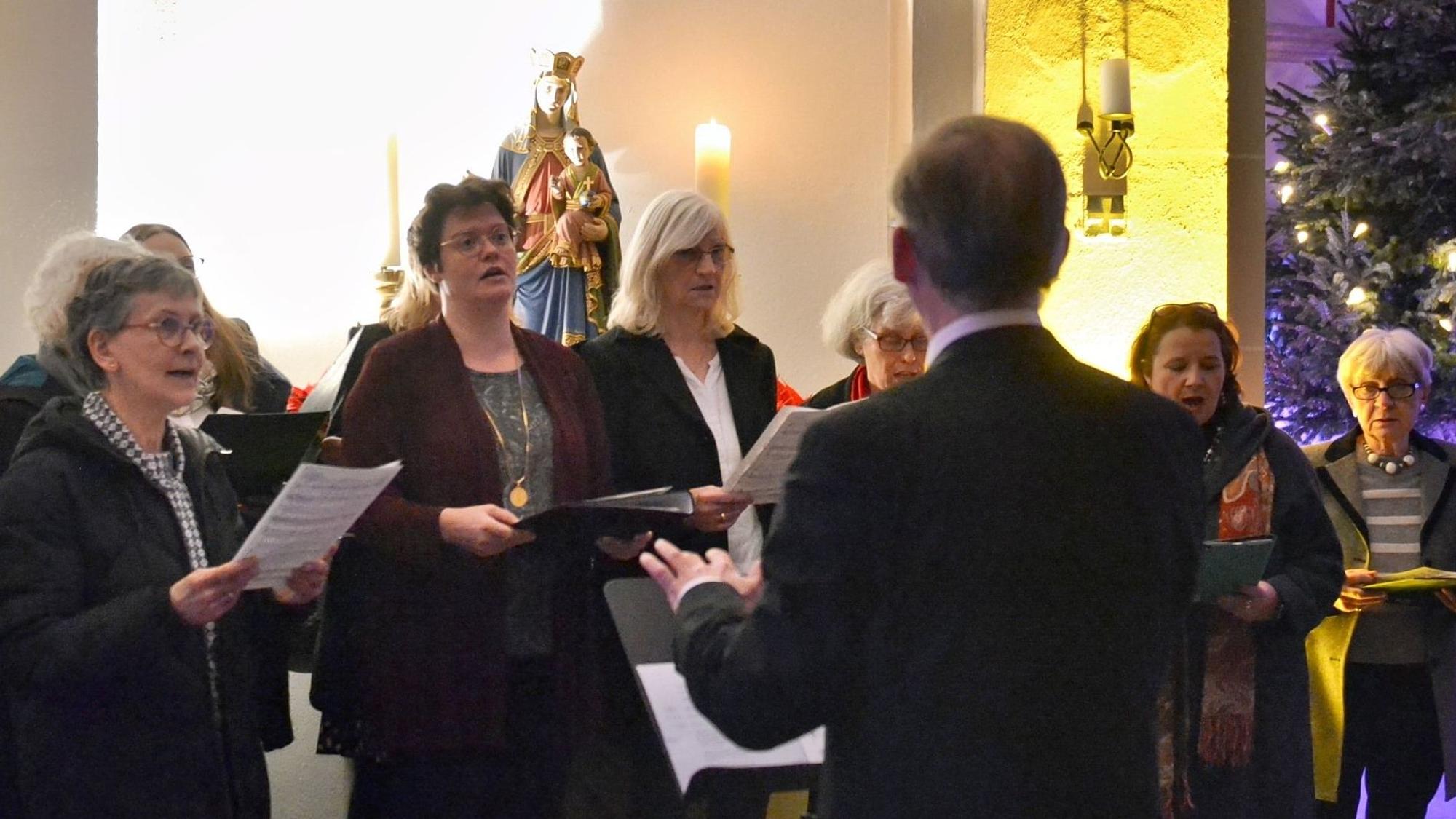 Mehrere Frauen halten Notenblätter in den Händen und singen, davor steht ein Dirigent, der nur von hinten zu sehen ist.