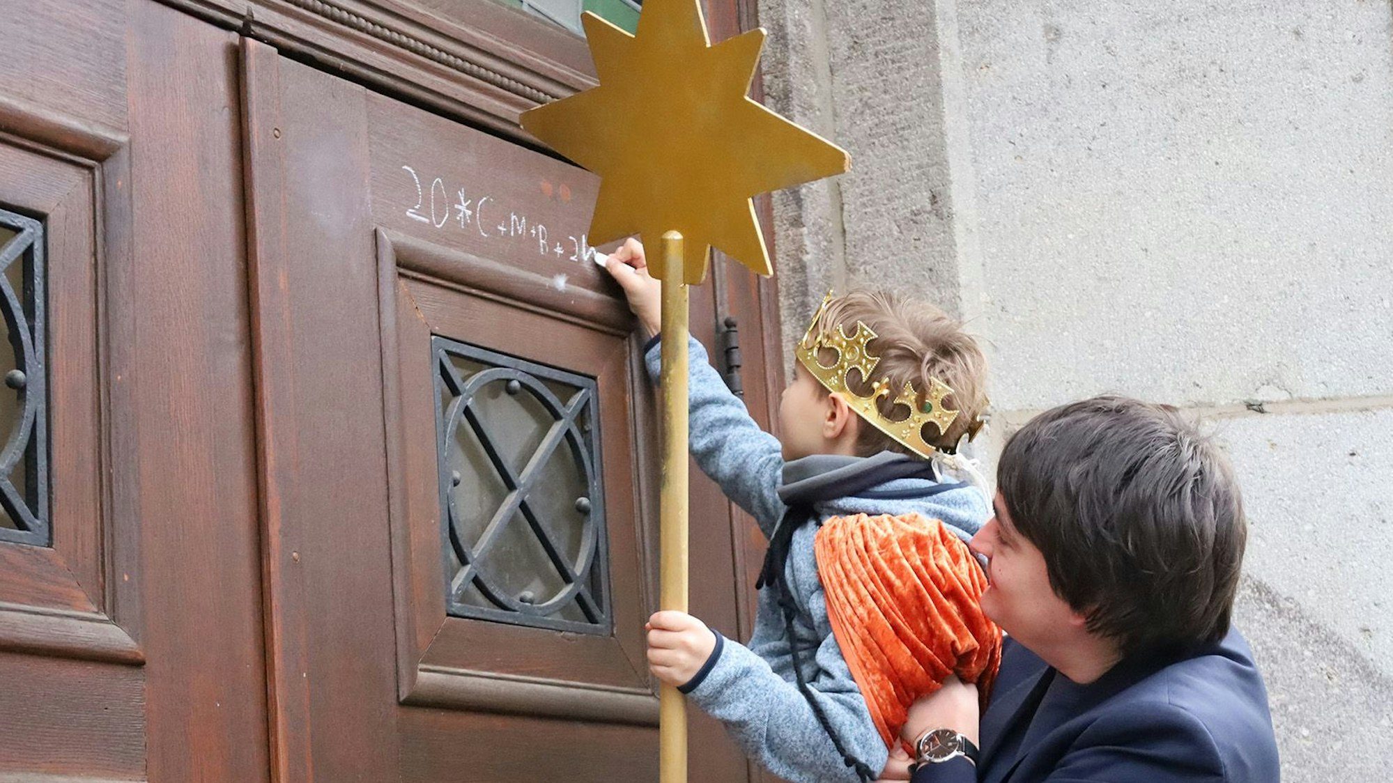 Ein Mann hebt ein Kind mit einer Krone und einem Stern hoch. Das Kind schreibt mit Kreide eine Zahl an eine Holztür.