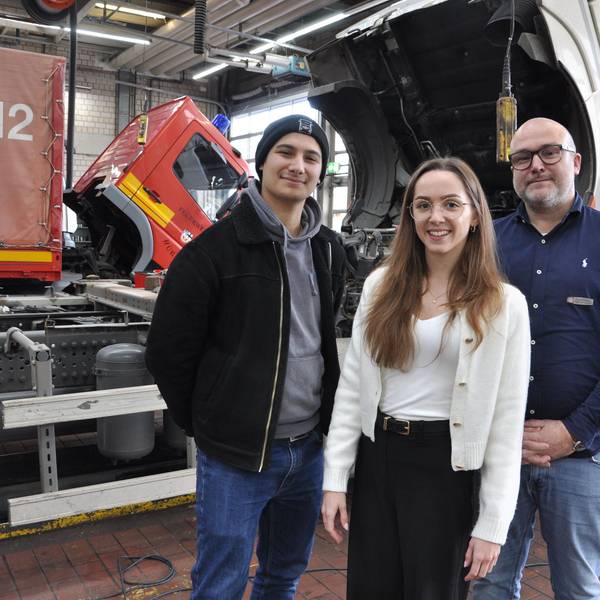 Auf dem Bild sind zwei Männer und eine junge Frau vor Lkws im Daimler Truck Nutzfahrzeugzentrums Mercedes-Benz in Frechen zu sehen.