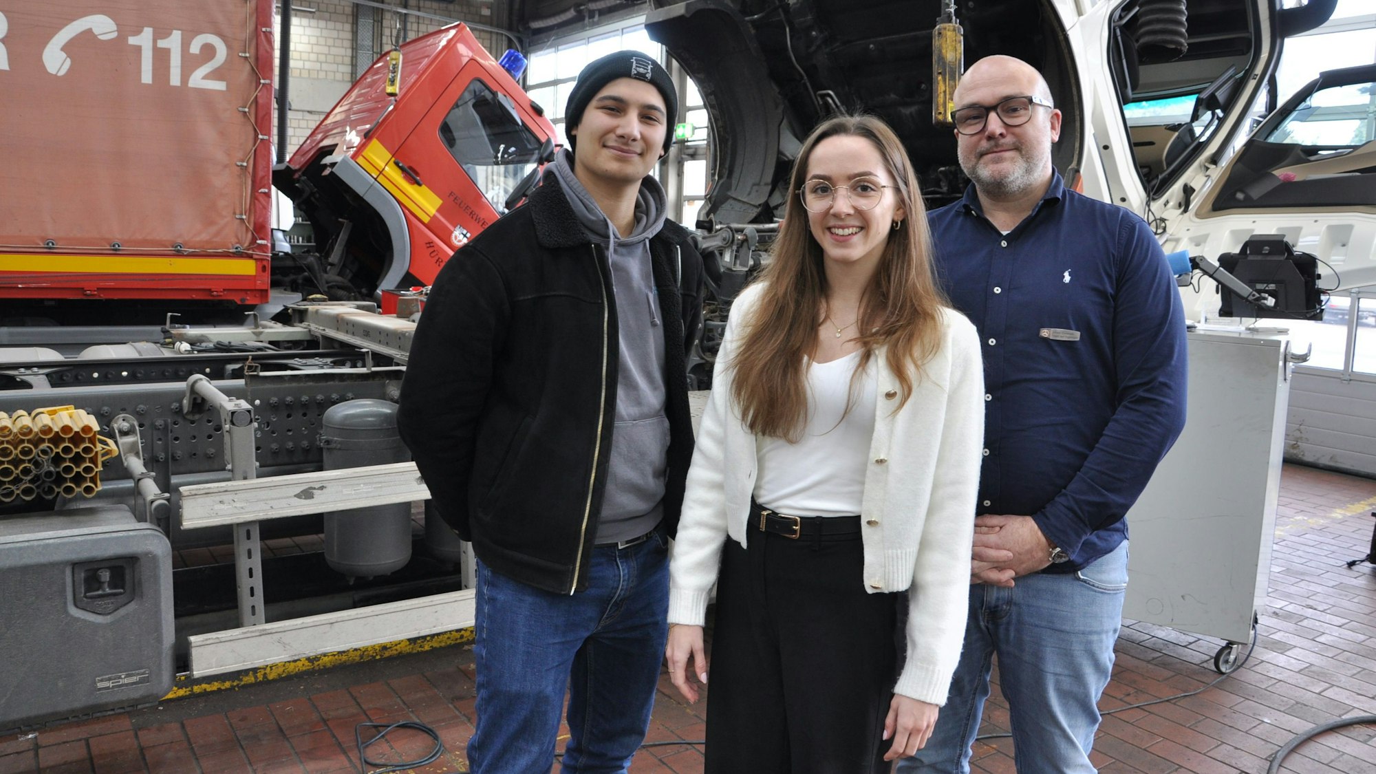 Auf dem Bild sind zwei Männer und eine junge Frau vor Lkws im Daimler Truck Nutzfahrzeugzentrums Mercedes-Benz in Frechen zu sehen.