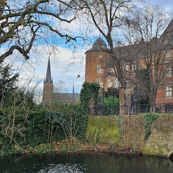 Zu sehen ist das Bedburger Schloss, ein Backsteinbau, sowie im Hintergrund die Kirche St. Lambertus.
