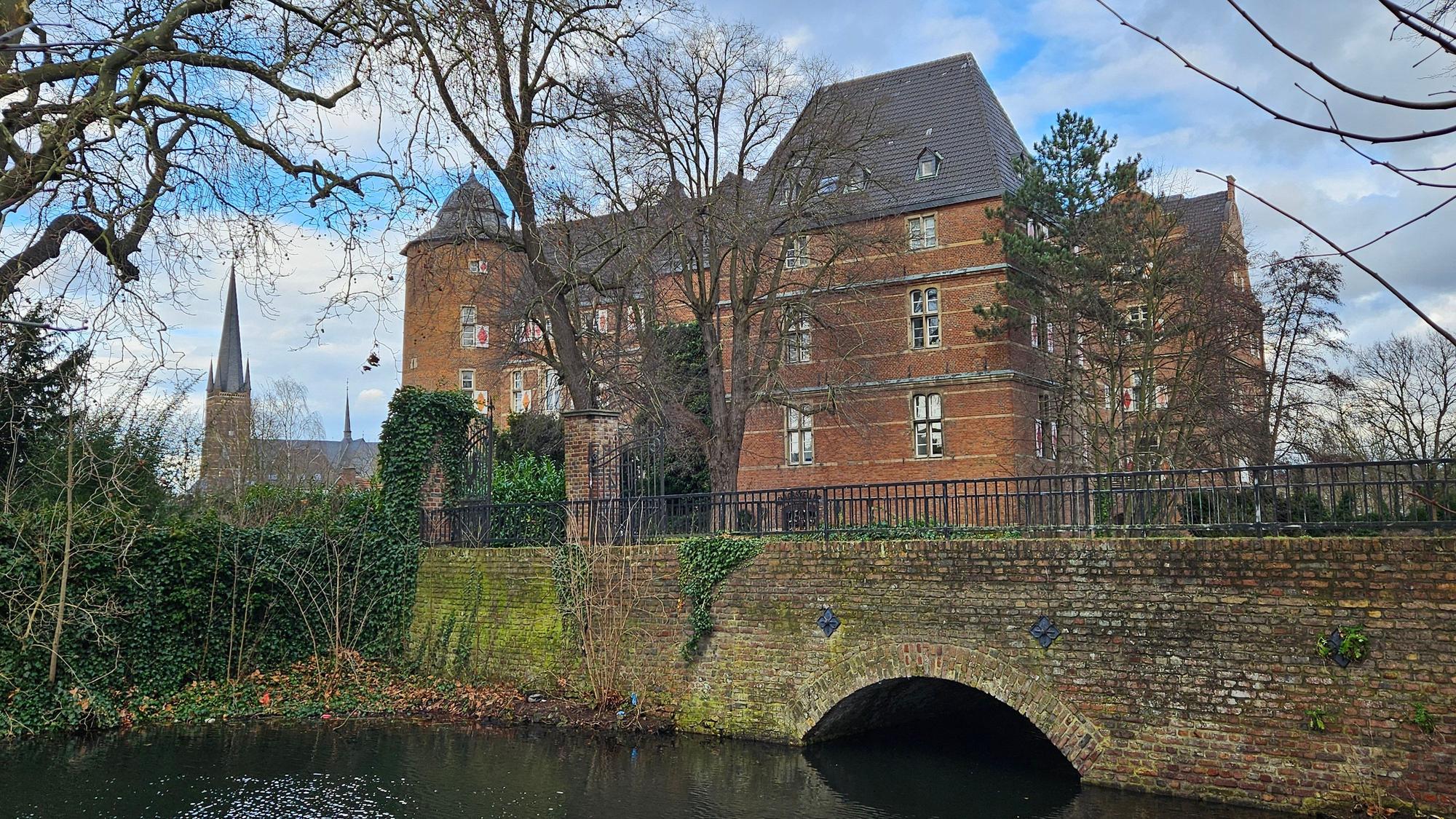 Zu sehen ist das Bedburger Schloss, ein Backsteinbau, sowie im Hintergrund die Kirche St. Lambertus.