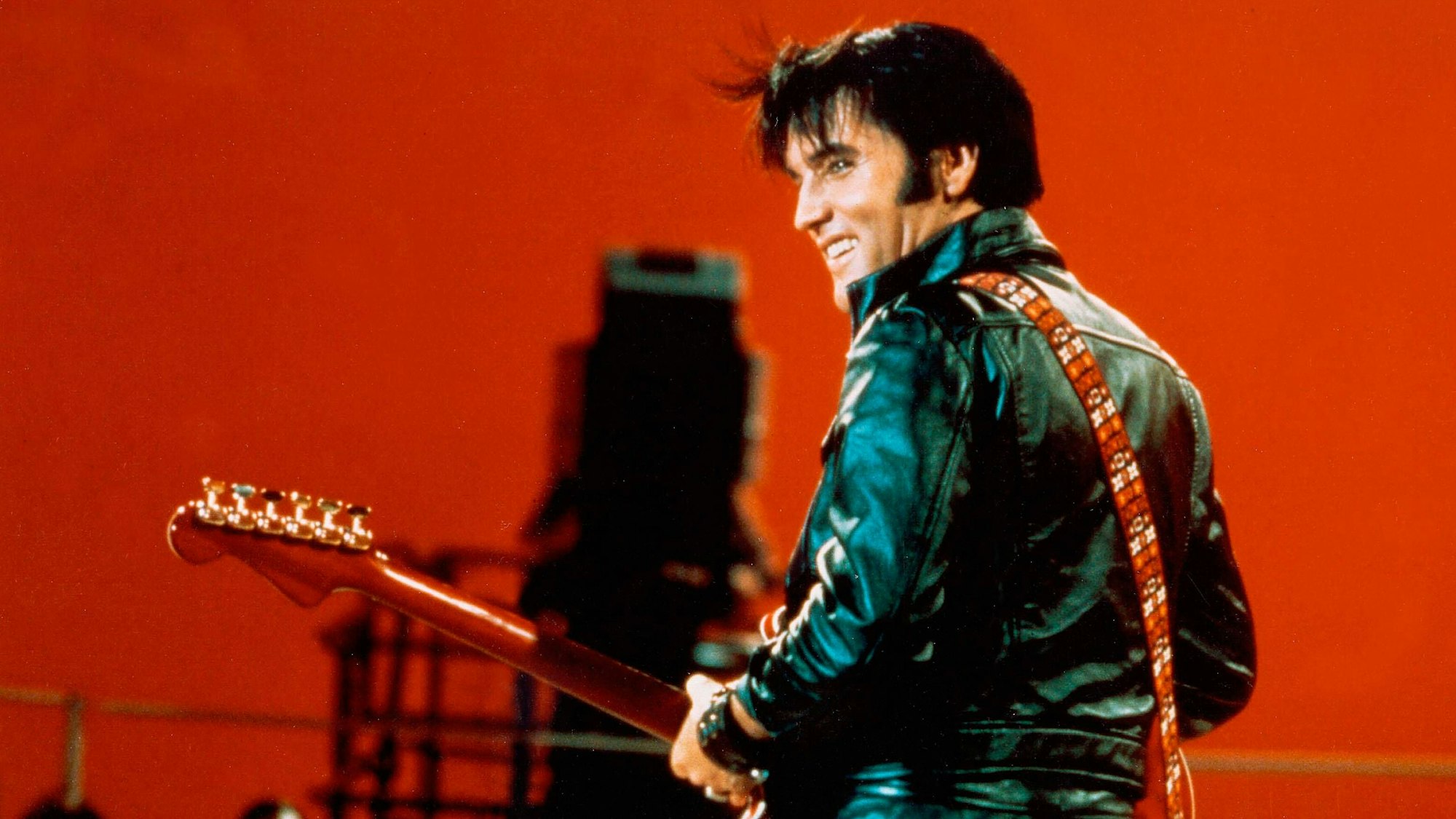Elvis hat in Rückansicht eine rote Gitarre umgeschnallt, er trägt eine schwarze Lederjacke und lacht.
