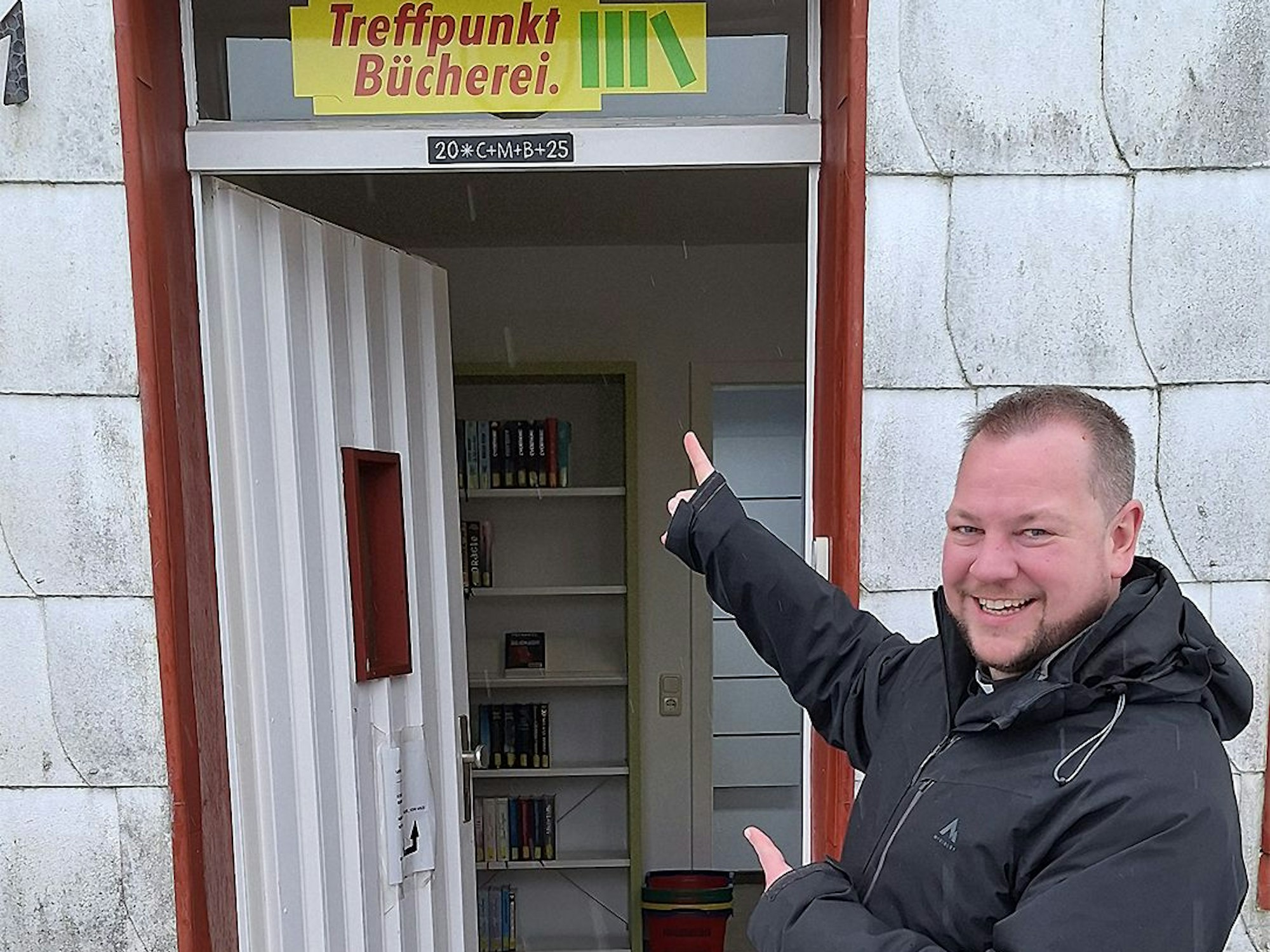 Pfarrer Thomas Schlütter steht vor der offenen Tür und zeigt auf das Schild der Bücherei.