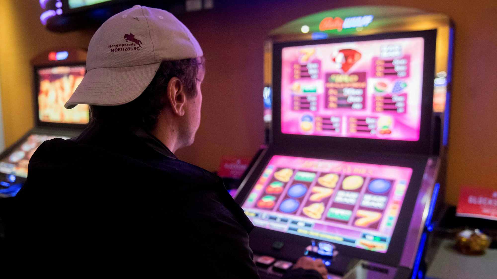 Ein Mann mit Baseballkappe sitzt mit dem Rücken zur Kamera an einem Spielautomaten.