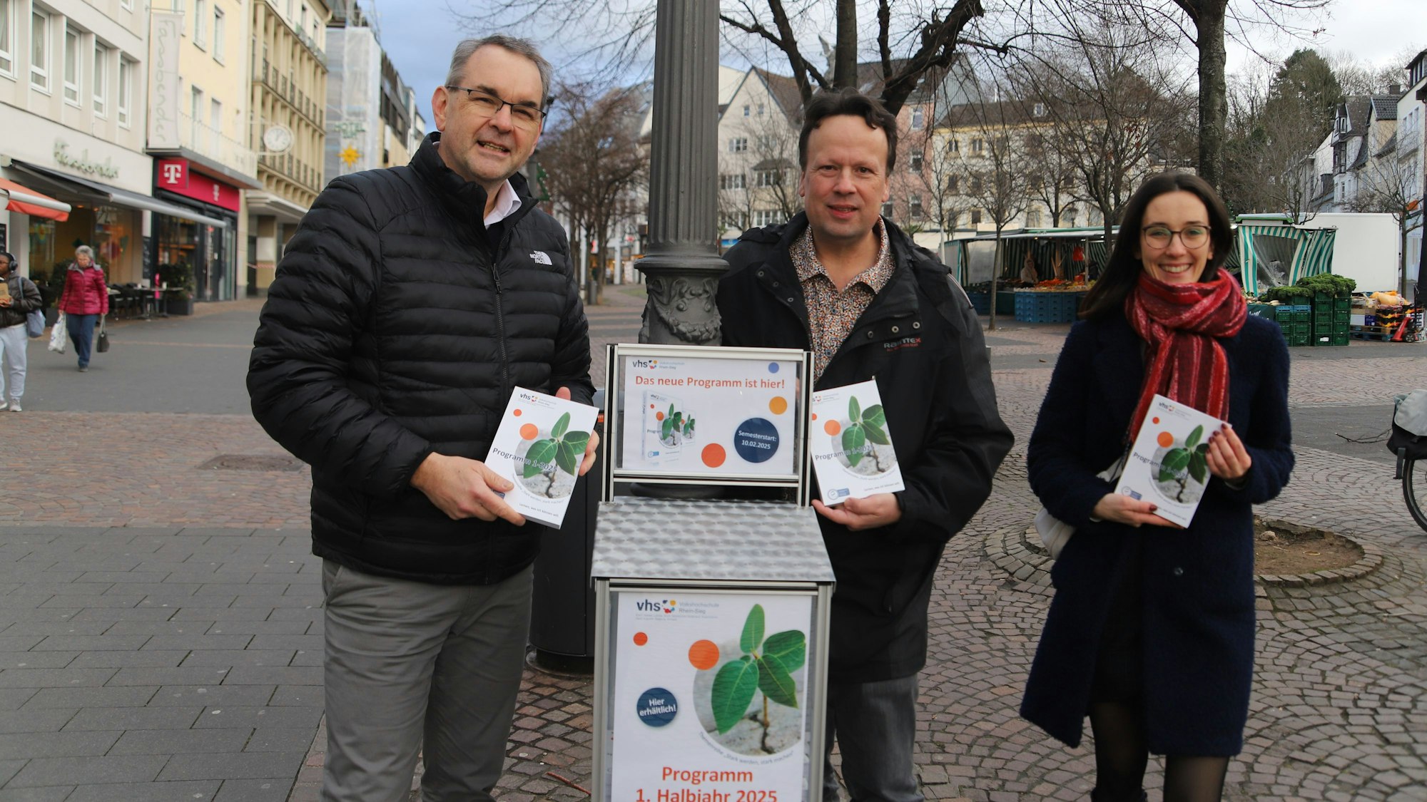 Zwei Männer und eine Frau stehen vor einem Kasten, aus dem Passanten das neue VHS-Programm entnehmen können.