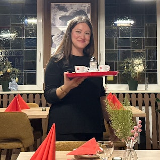 Eine Frau mit dunklen Haaren hält ein Tablett in der Hand, auf dem Espresso-Tassen stehen. Sie steht im Gastraum des Restaurants Buon Cibo in Köln-Zündorf.