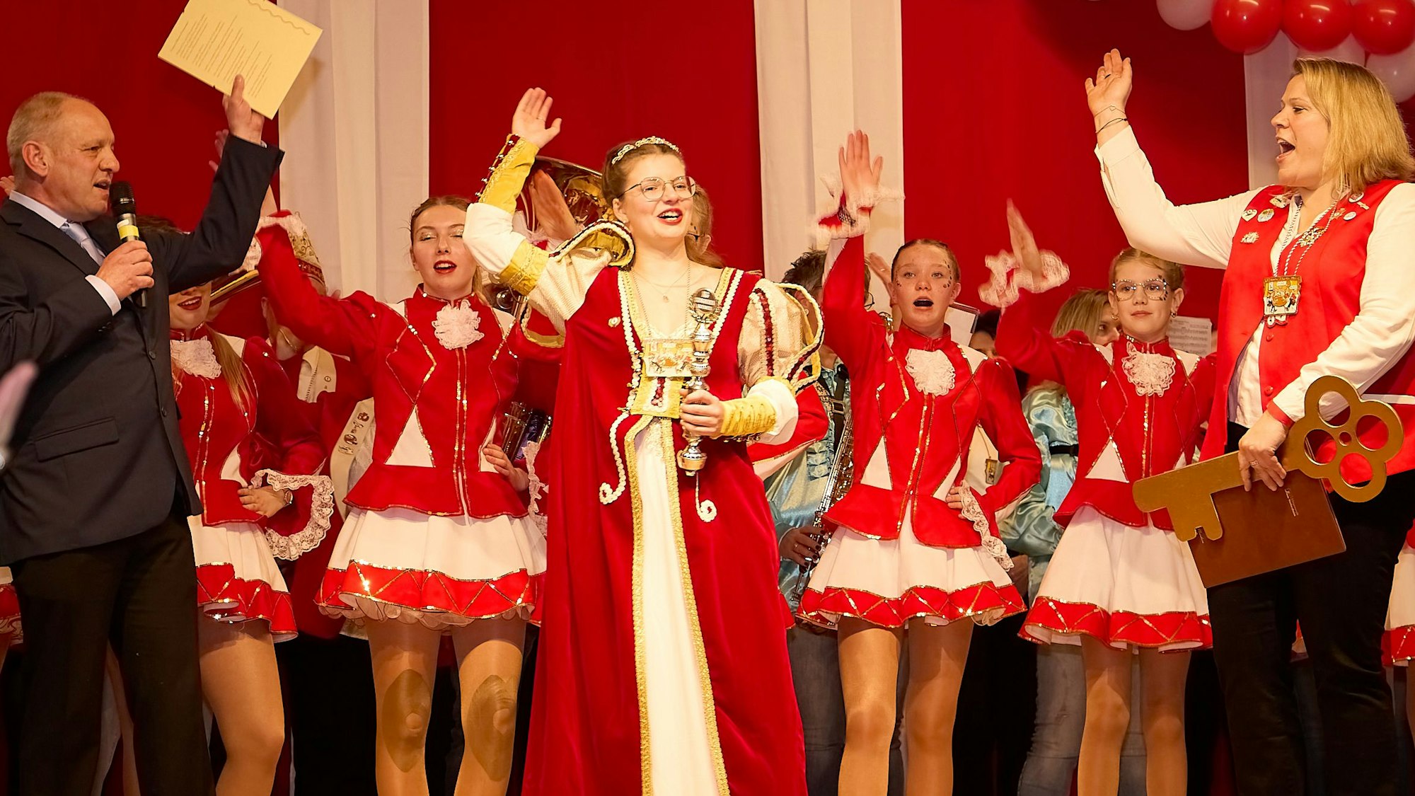 Karnevals-Prinzessin Ophelia I. steht im rot-weißen Ornat zusammen mit den Tänzerinnen einer Garde und weiteren Personen auf der Bühne; alle heben die Hand Karnevalsgruß.