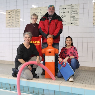 Eine Gruppe Männer und Frauen, unter anderem von der DLRG, stehen mit einer Trainingspuppe am Beckenrand eines Hallenbads.