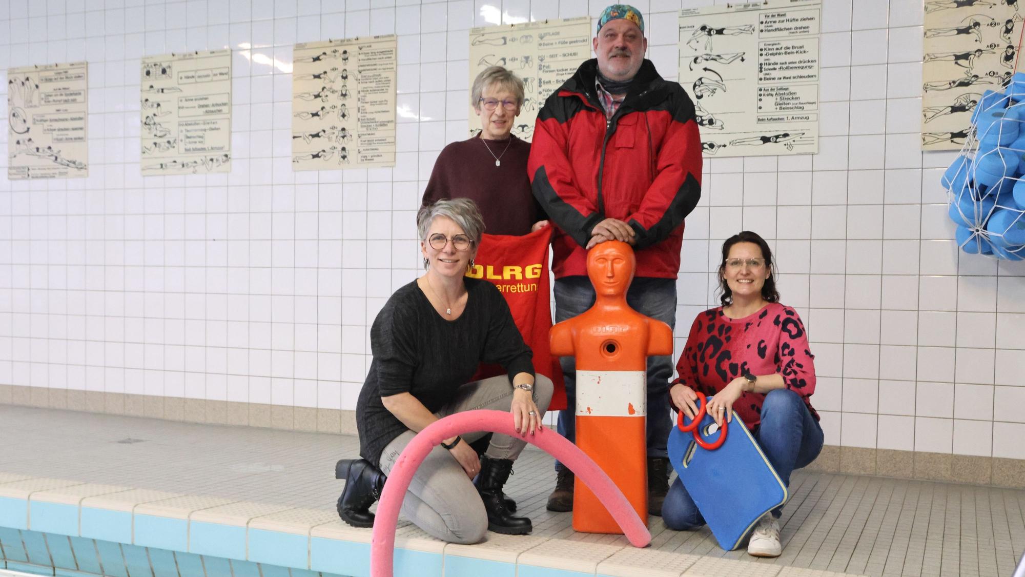 Eine Gruppe Männer und Frauen, unter anderem von der DLRG, stehen mit einer Trainingspuppe am Beckenrand eines Hallenbads.