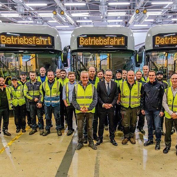 Auf dem Foto sind Männer in Anzügen und andere in gelben Warnwesten vor Bussen zu sehen.