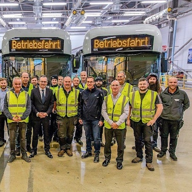 Auf dem Foto sind Männer in Anzügen und andere in gelben Warnwesten vor Bussen zu sehen.