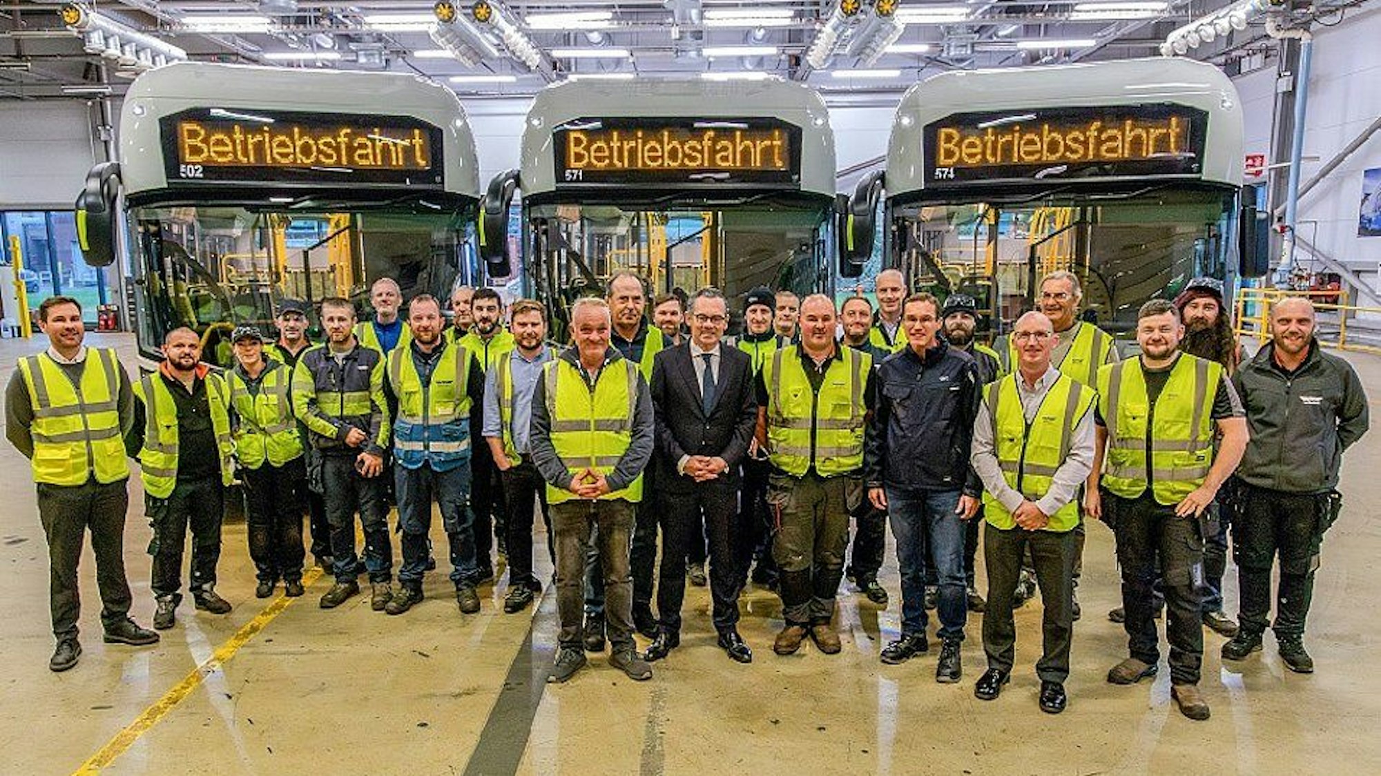 Auf dem Foto sind Männer in Anzügen und andere in gelben Warnwesten vor Bussen zu sehen.
