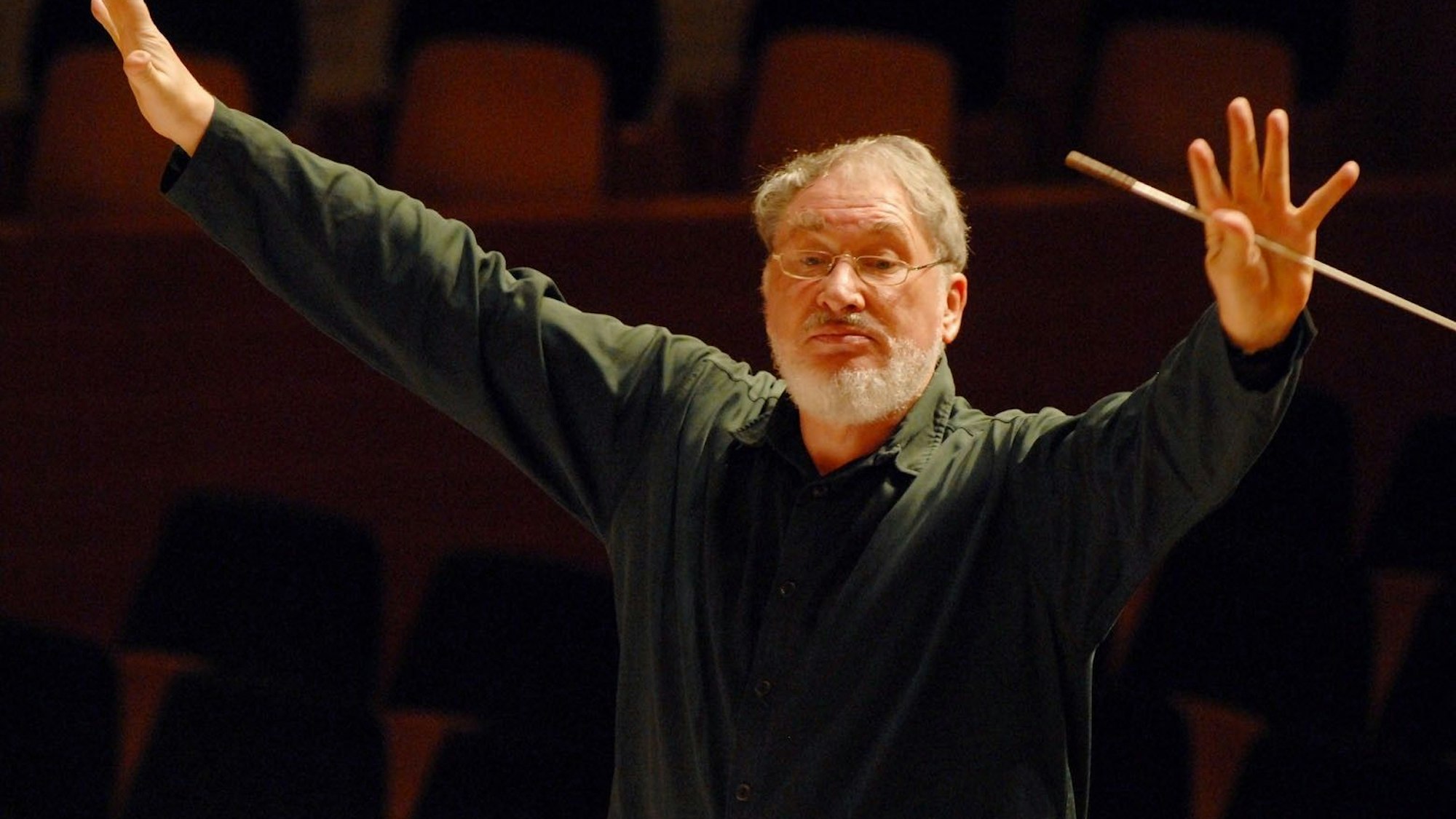 HK Gruber steht mit ausgebreiteten Armen auf einer Bühne.