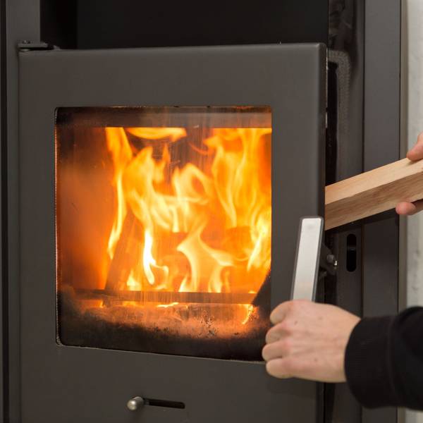 Ein Mann legt in seiner Wohnung ein Stück Holz in einen Kaminofen, in dem ein Feuer brennt.