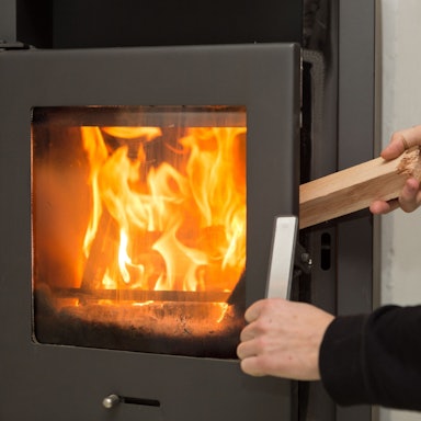 Ein Mann legt in seiner Wohnung ein Stück Holz in einen Kaminofen, in dem ein Feuer brennt.