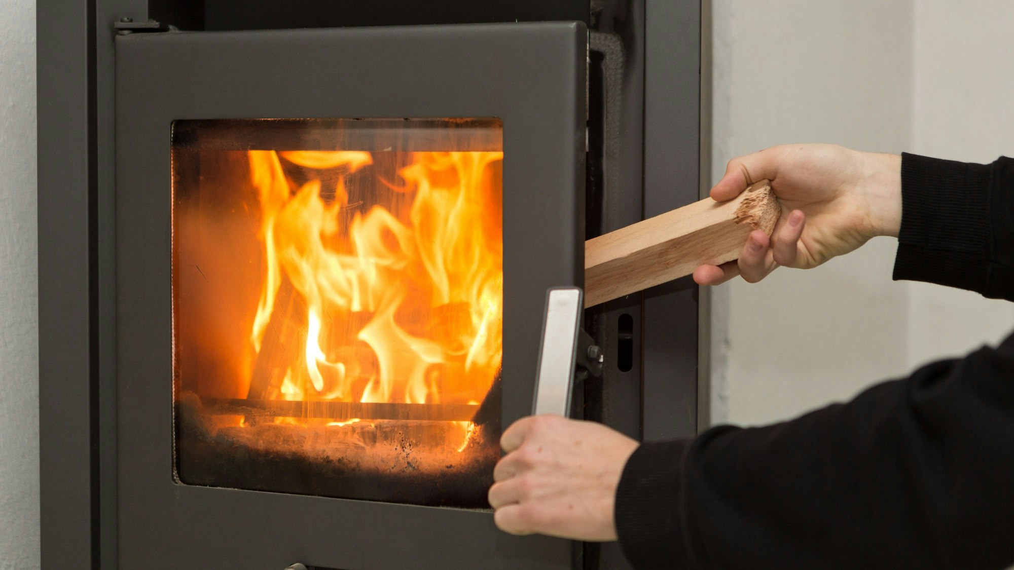 Ein Mann legt in seiner Wohnung ein Stück Holz in einen Kaminofen, in dem ein Feuer brennt.