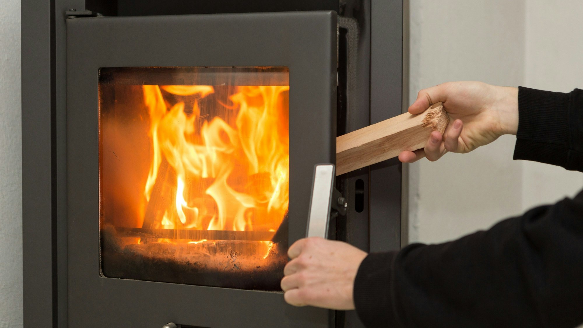 Ein Mann legt in seiner Wohnung ein Stück Holz in einen Kaminofen, in dem ein Feuer brennt.