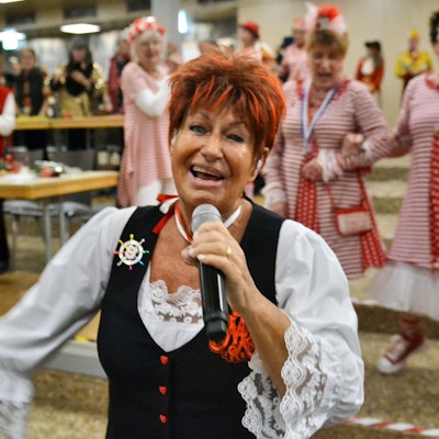 Marita Köllner singt und strahlt in die Kamera. Im Hintergrund schunkeln kostümierte Besucher.
