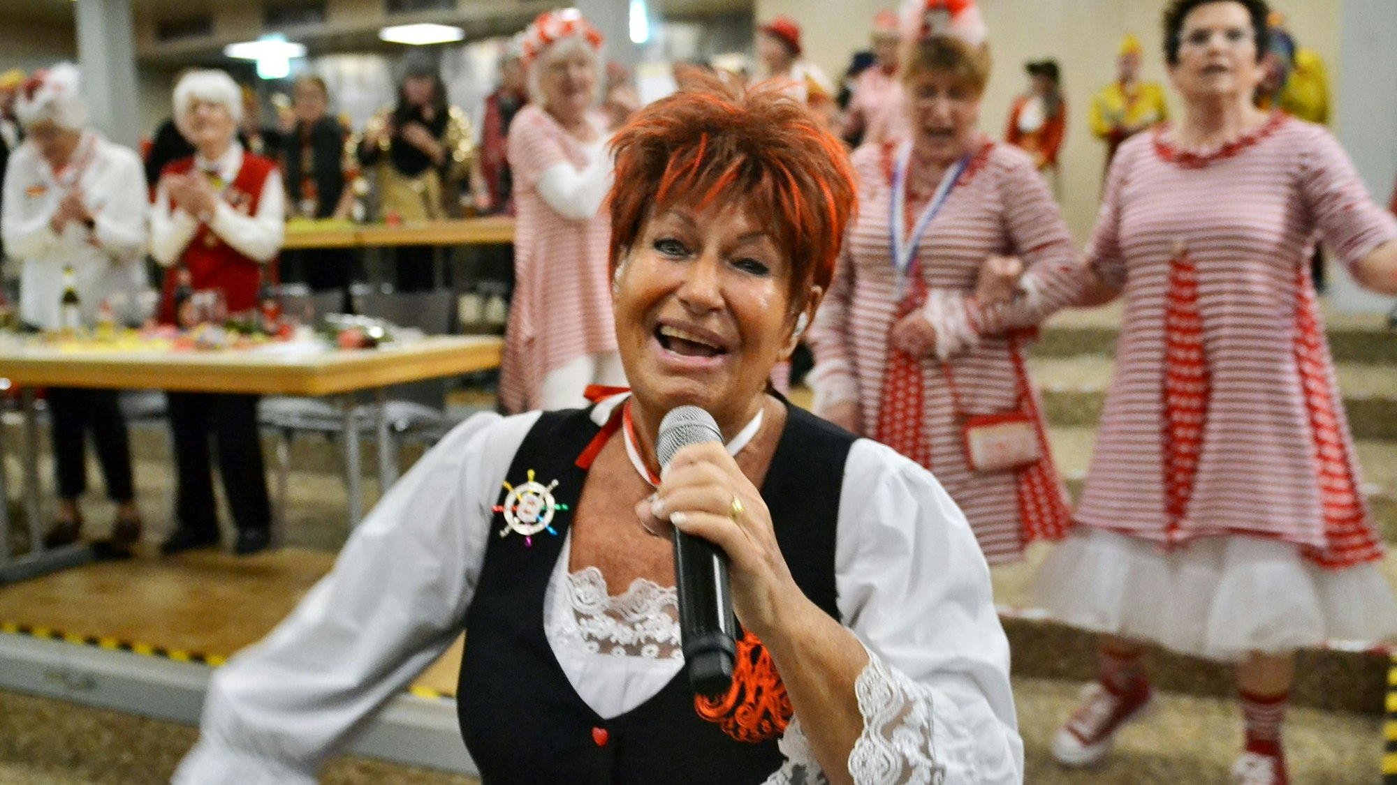 Marita Köllner singt und strahlt in die Kamera. Im Hintergrund schunkeln kostümierte Besucher.