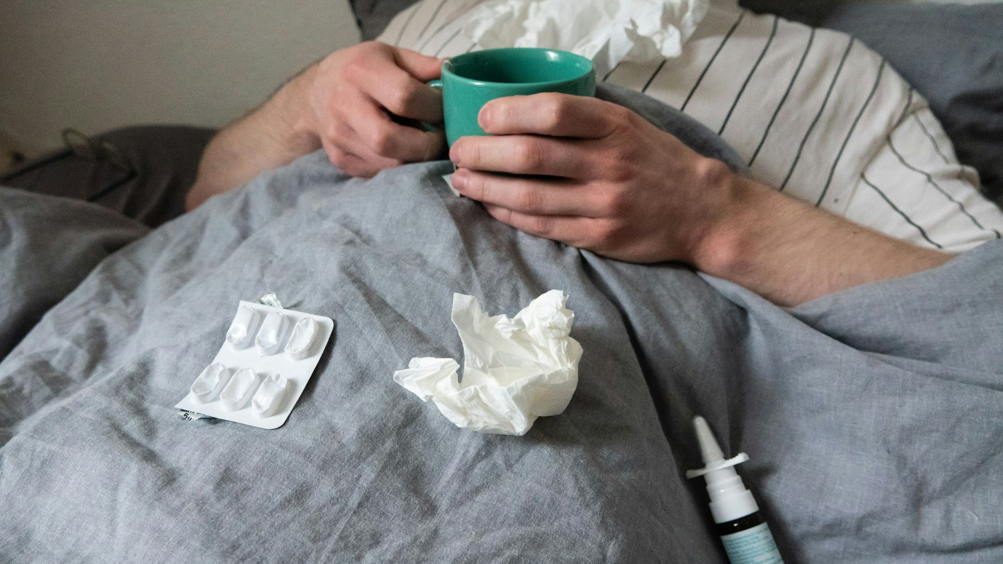 ILLUSTRATION - Ein Mann liegt mit Taschentüchern, Teetasse, Nasenspray und Tabletten im Bett (gestellte Szene). Derzeit bewegt sich die Rate akuter Atemwegserkrankungen in der Bevölkerung weiter auf einem relativ hohen Niveau im Vergleich zu früheren Jahren. +++ dpa-Bildfunk +++