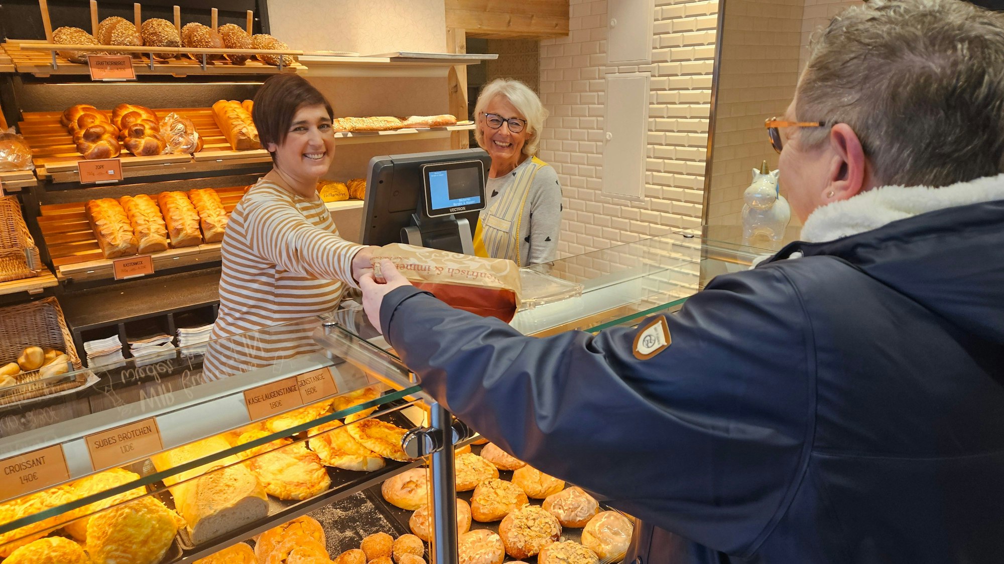 Zu sehen sind zwei Frauen hinter der Verkaufstheke einer Bäckerei, eine der beiden reicht einer Kundin eine Tüte mit Brötchen.